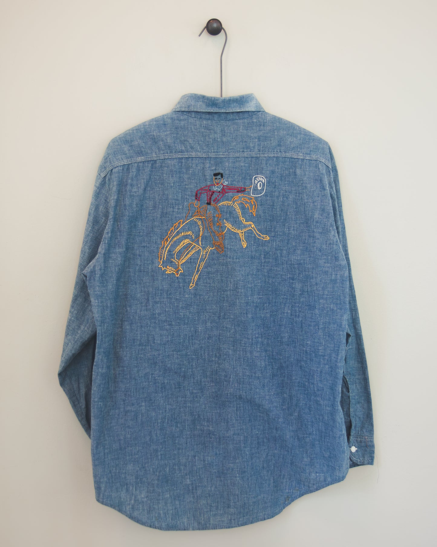 A blue denim long-sleeve button-up shirt hanging on a black hanger against a plain off-white wall. The back of the shirt features a colorful embroidered graphic of a cowboy riding a bucking horse.