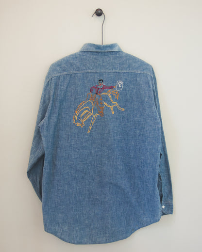 A blue denim long-sleeve button-up shirt hanging on a black hanger against a plain off-white wall. The back of the shirt features a colorful embroidered graphic of a cowboy riding a bucking horse.