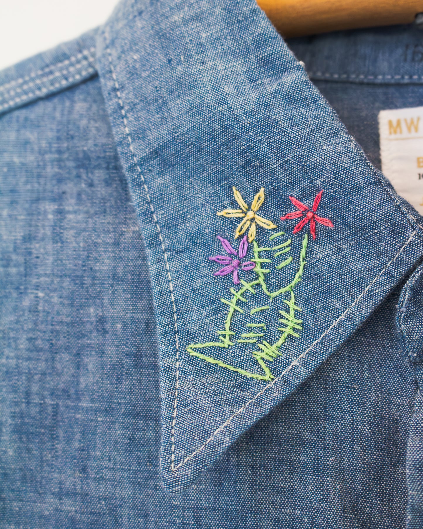 Close-up of a blue denim shirt collar with colorful floral embroidery on the point, showing yellow, purple, and red flowers with green stems and leaves.