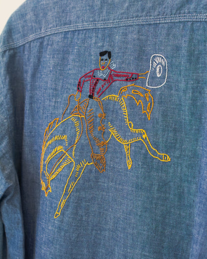 Close-up of a blue denim shirt featuring a colorful embroidered graphic of a cowboy riding a bucking bronco, with the cowboy holding a white hat.
