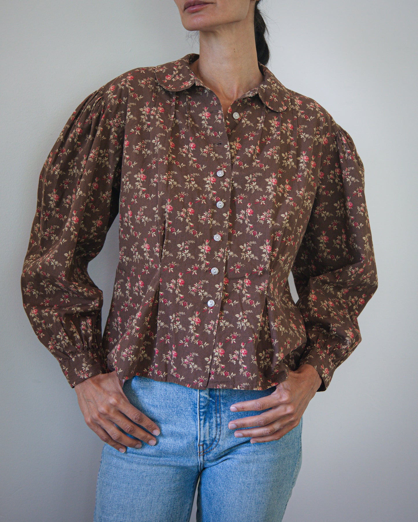 A woman wearing a vintage-style brown button-up blouse with a dense small floral print, paired with light blue jeans, photographed against a plain light gray wall.