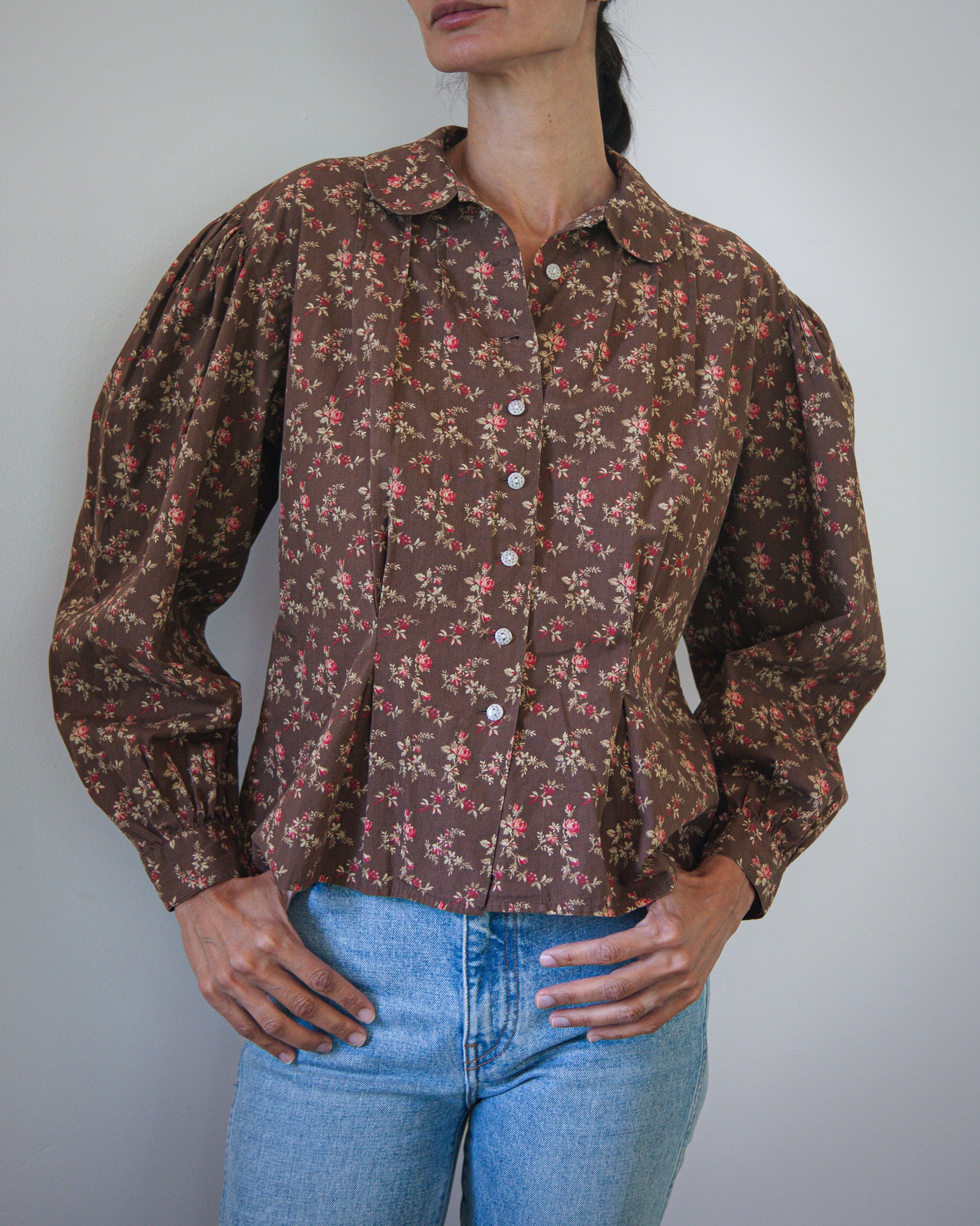 A woman wearing a vintage-style brown button-up blouse with a dense small floral print, paired with light blue jeans, photographed against a plain light gray wall.