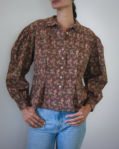 A woman wearing a vintage-style brown button-up blouse with a dense small floral print, paired with light blue jeans, photographed against a plain light gray wall.
