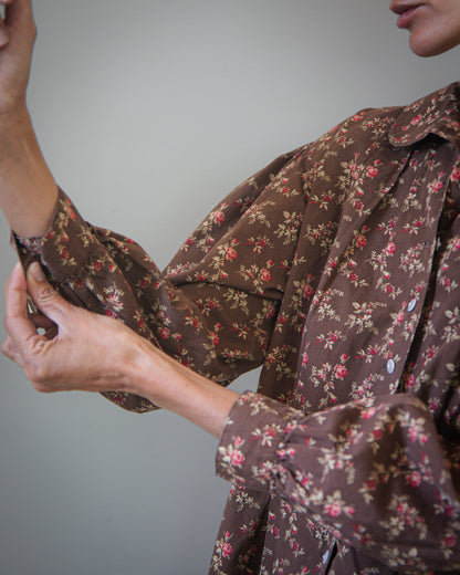 A close-up of a person wearing a brown long-sleeve button-up shirt with a dense pink and beige floral print, adjusting the cuff of the sleeve against a plain gray background.