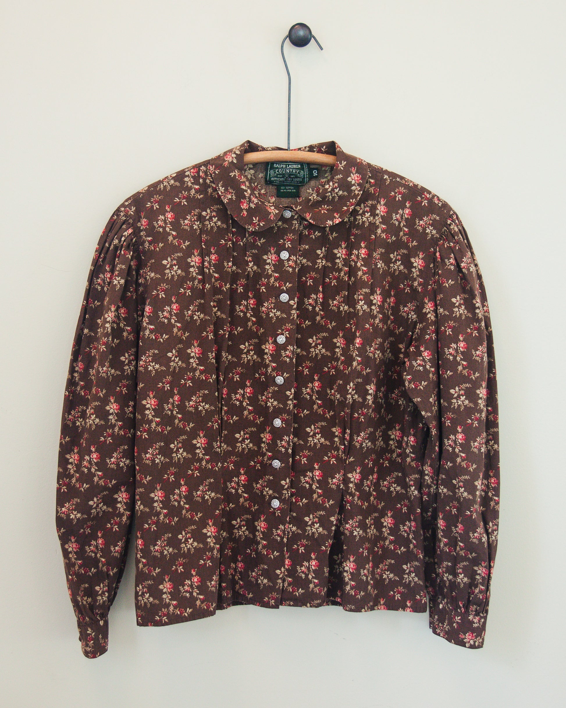 A vintage floral print button-up blouse hanging on a wooden hanger against a plain off-white wall. The shirt features a dark brown base with a dense pattern of small pink, red, and beige flowers.