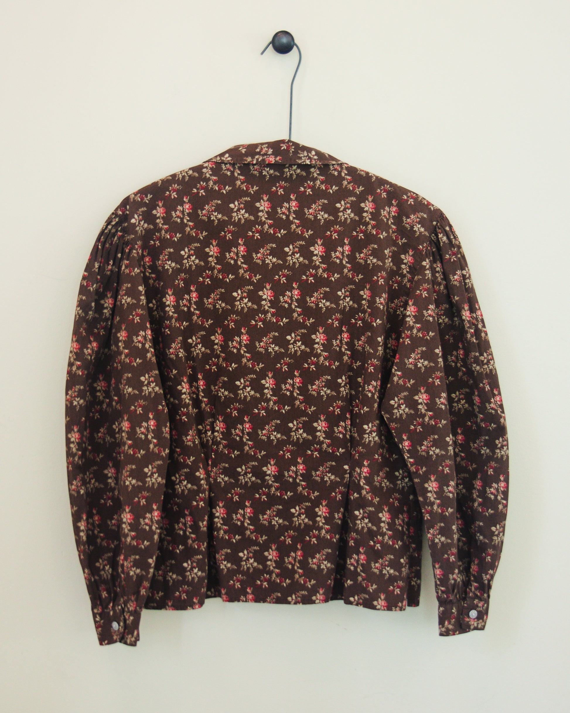 Back of a long-sleeve button-up blouse with a dense small floral print, hanging on a black hanger against a plain off-white wall.