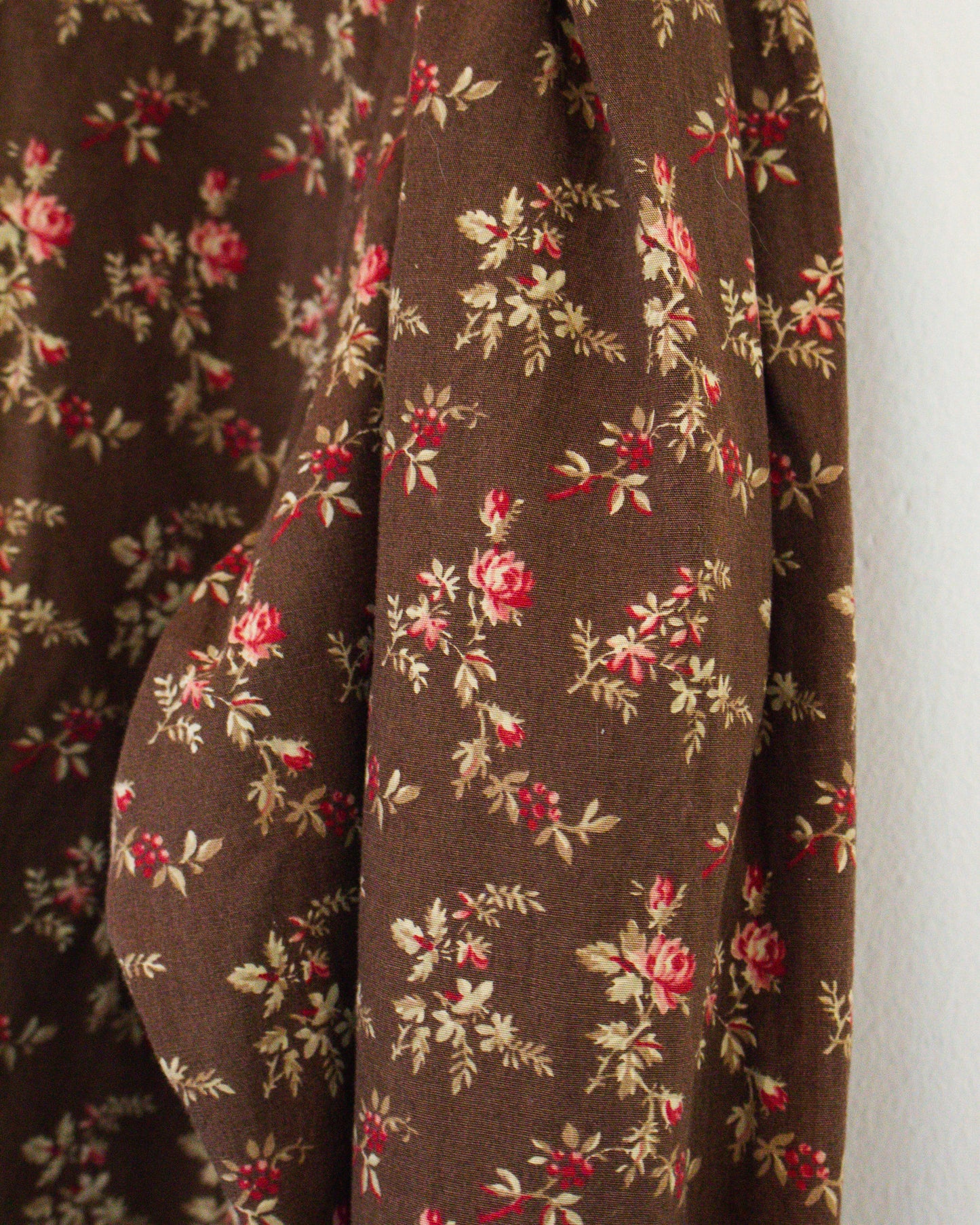 A close-up of brown fabric with a vintage-style floral print featuring red flowers and beige leaves against a plain white wall.