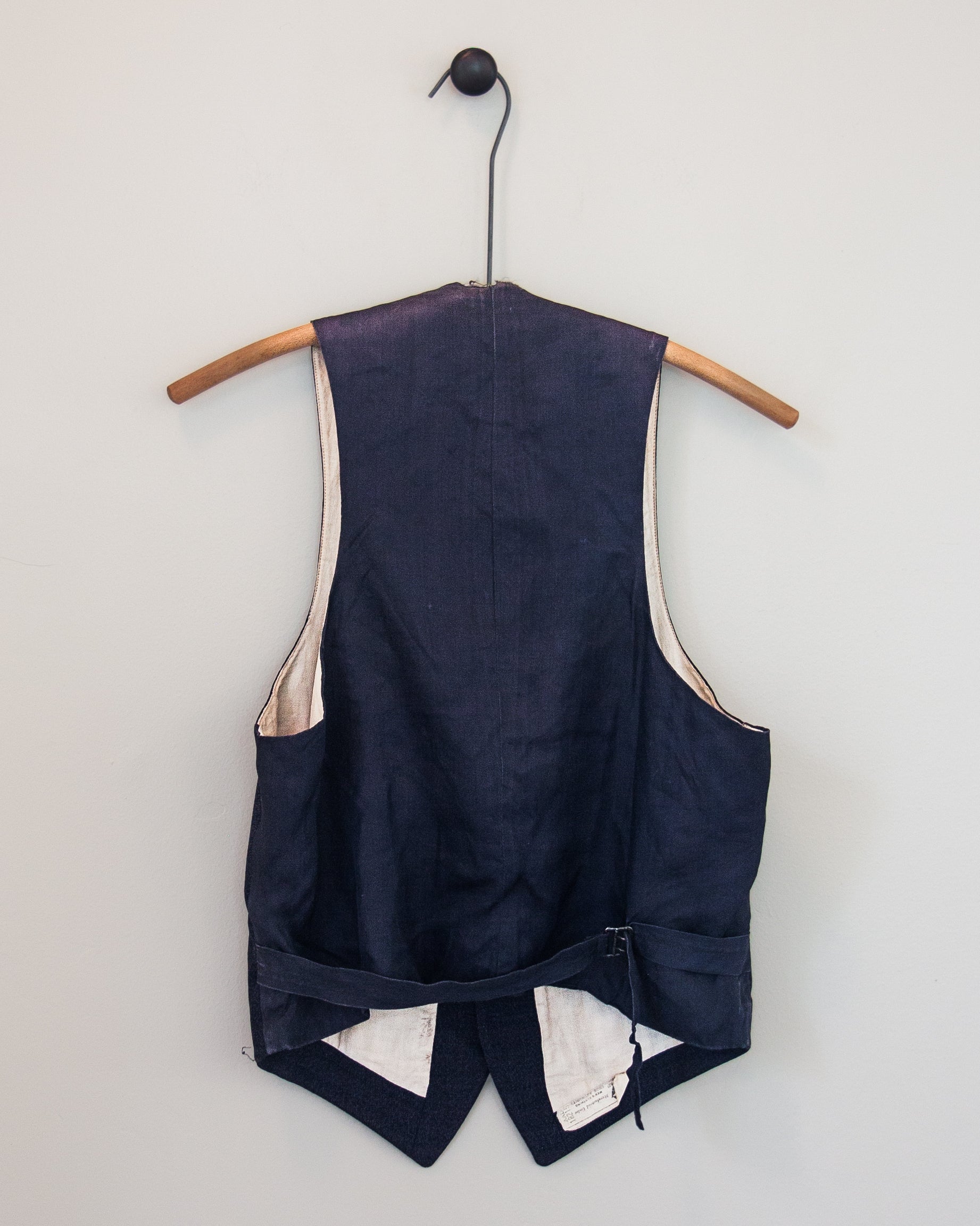 A navy blue vest shown from the back, hanging on a wooden hanger against a light gray wall. The vest features cream-colored lining and a back cinch closure at the waist.