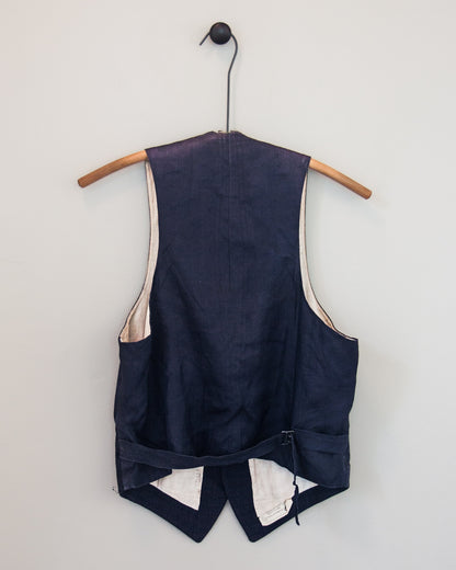 A navy blue vest shown from the back, hanging on a wooden hanger against a light gray wall. The vest features cream-colored lining and a back cinch closure at the waist.