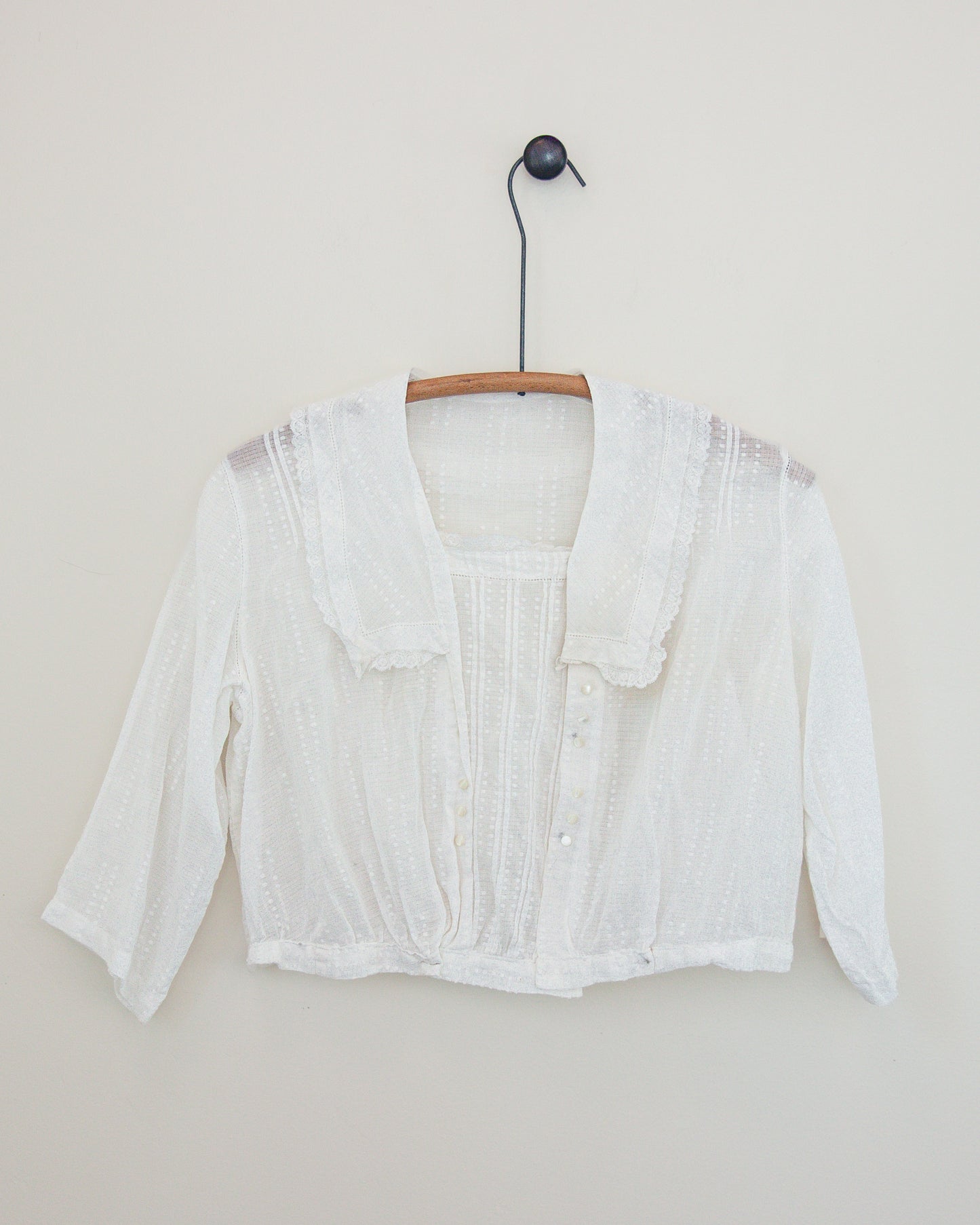 A white textured lace and cotton top with a ruffled collar and button details, hanging on a wooden hanger against a plain beige wall.