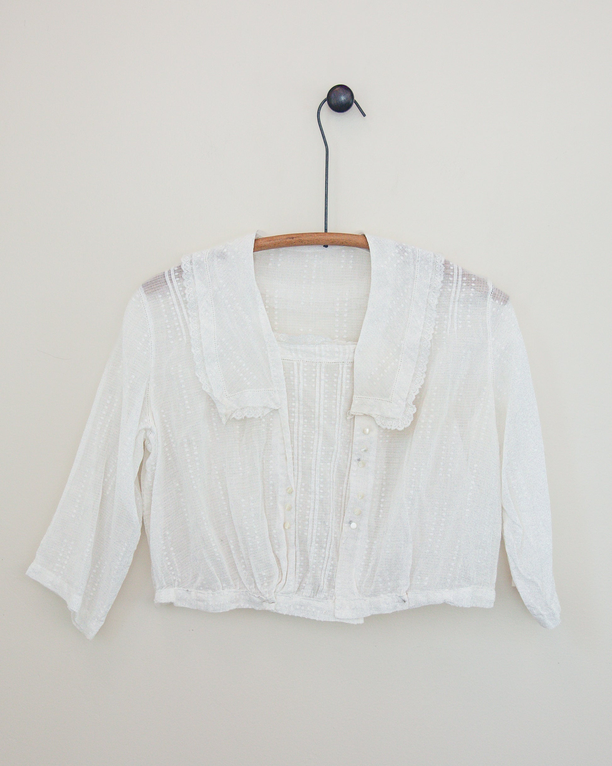 A white textured lace and cotton top with a ruffled collar and button details, hanging on a wooden hanger against a plain beige wall.