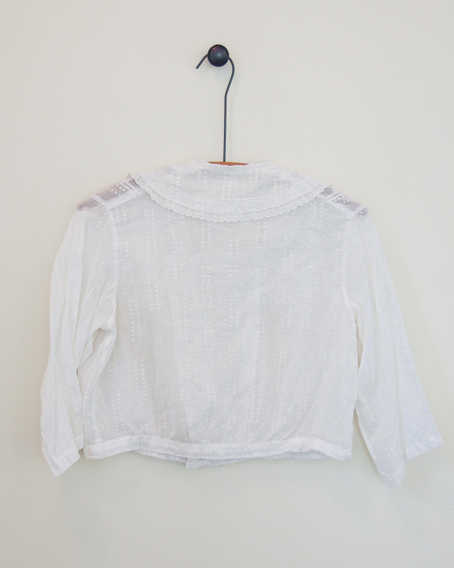 Back of a white blouse with lace shoulder details and a scalloped neckline, hanging on a black hanger against a plain off-white wall.