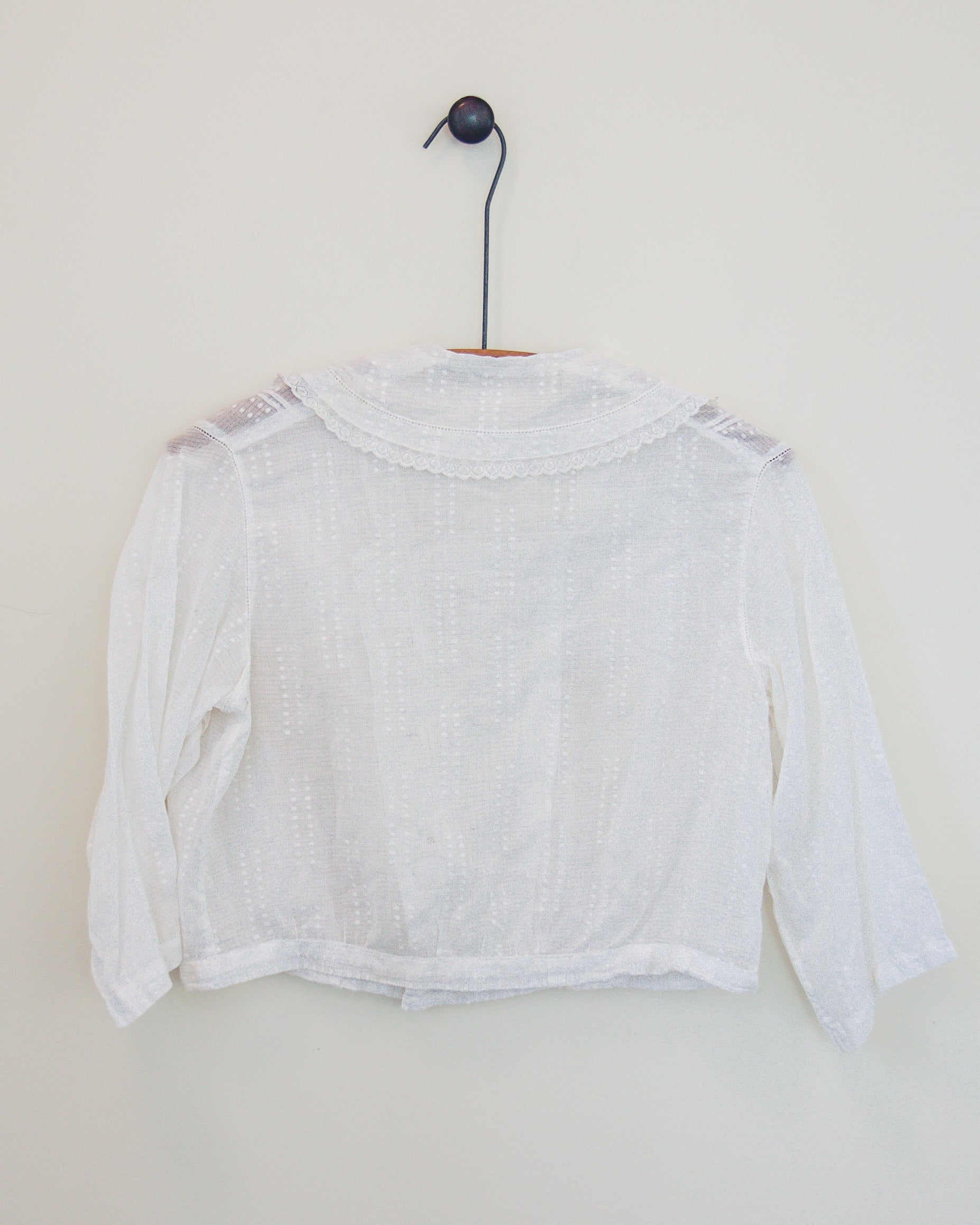 Back of a white blouse with lace shoulder details and a scalloped neckline, hanging on a black hanger against a plain off-white wall.