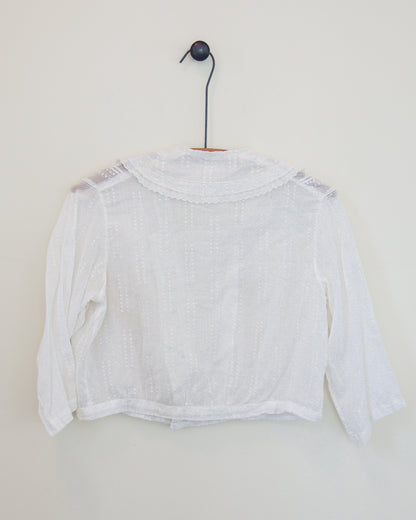 Back of a white blouse with lace shoulder details and a scalloped neckline, hanging on a black hanger against a plain off-white wall.