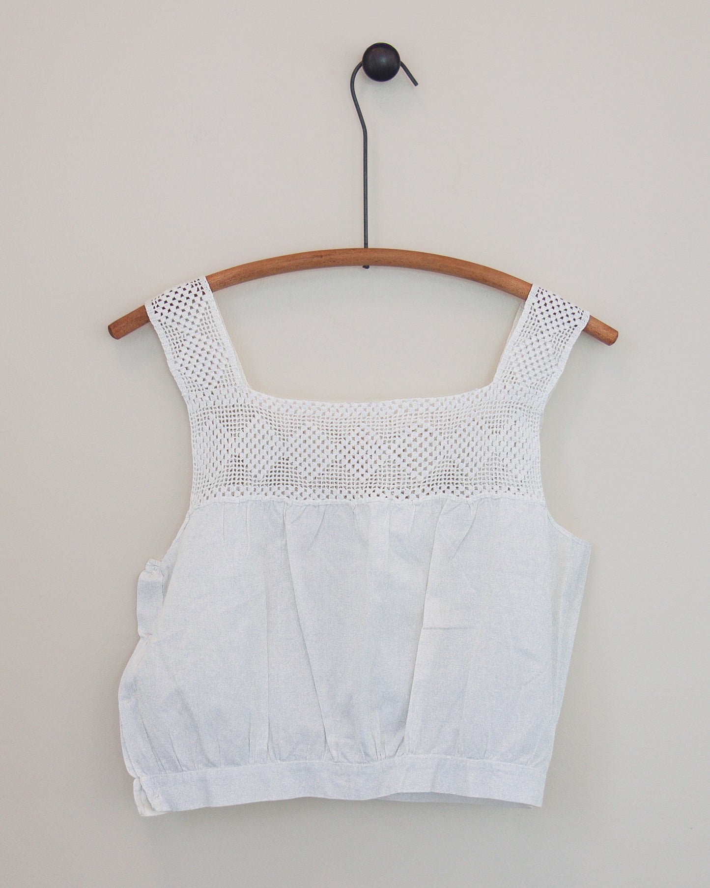 A white sleeveless top with a textured crochet yoke and square neckline, hanging on a wooden hanger against a plain light gray wall.