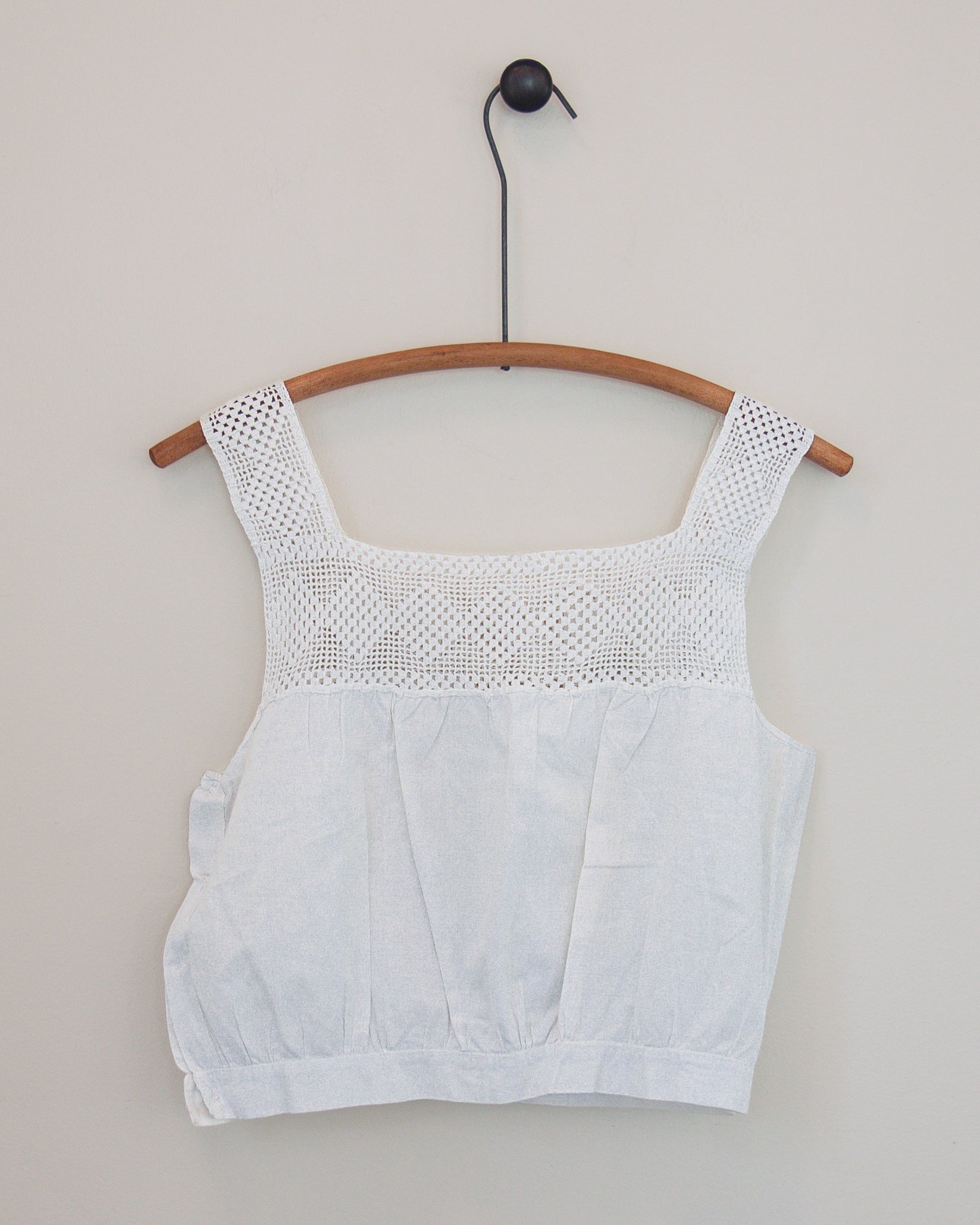 A white sleeveless top with a textured crochet yoke and square neckline, hanging on a wooden hanger against a plain light gray wall.