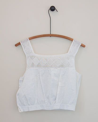 A white sleeveless top with a textured crochet yoke and square neckline, hanging on a wooden hanger against a plain light gray wall.