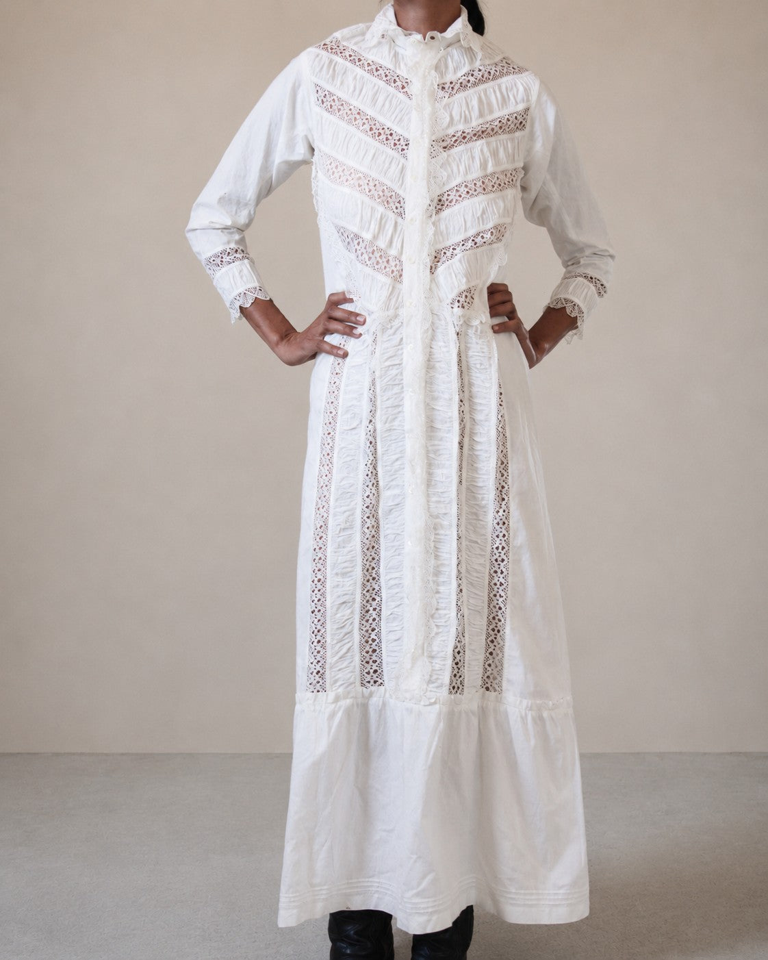 A long white embroidered nightgown with vertical lace panels and ruffled detailing, worn by a woman standing against a plain beige studio background.