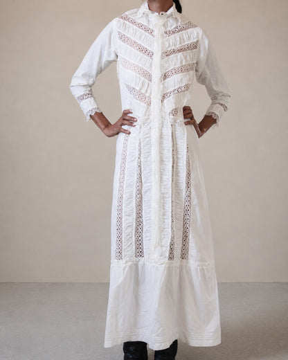 A long white embroidered nightgown with vertical lace panels and ruffled detailing, worn by a woman standing against a plain beige studio background.