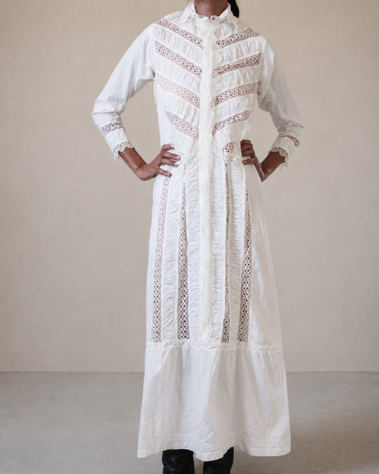 A long white embroidered nightgown with vertical lace panels and ruffled detailing, worn by a woman standing against a plain beige studio background.