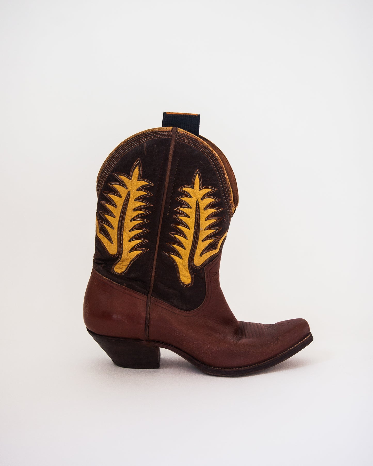 A single brown leather cowboy boot with dark brown shaft panels featuring yellow embroidered feather designs, shown from a side profile on a white background.