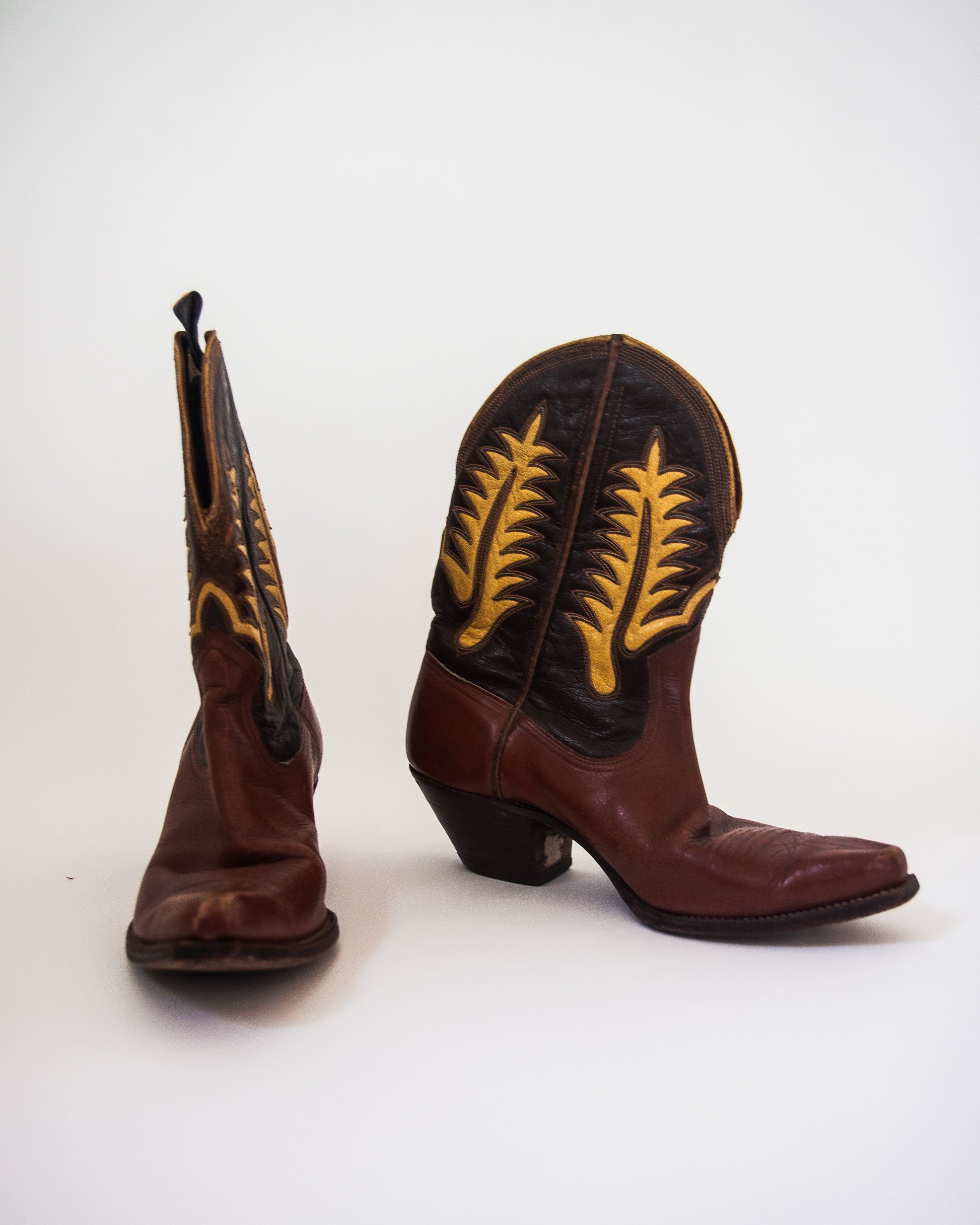 A pair of brown leather cowboy boots with yellow embroidered leaf patterns on the shafts, shown against a plain white background.