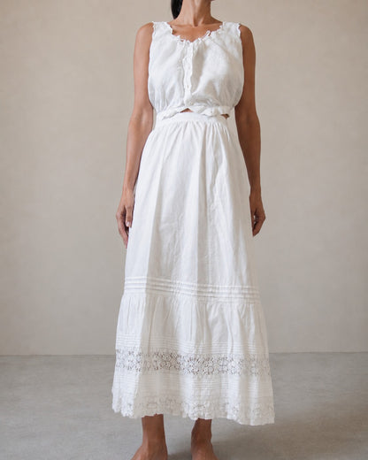 A woman modeling a sleeveless white top with a white cotton skirt that has an embroidered lace hem against a neutral beige studio background.