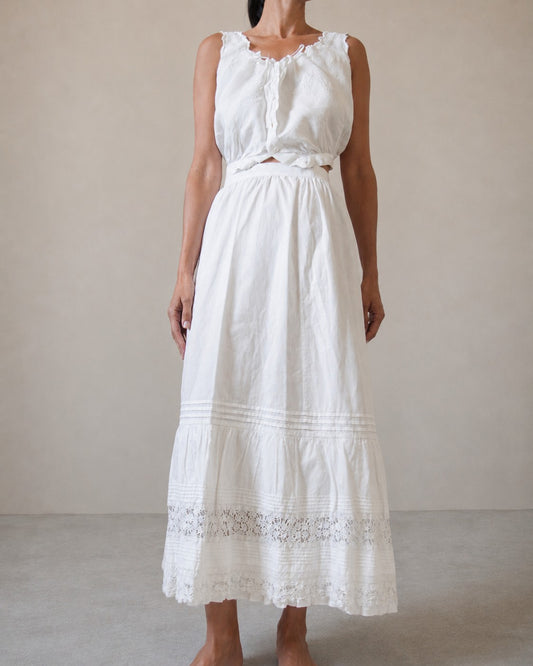 A woman modeling a sleeveless white top with a white cotton skirt that has an embroidered lace hem against a neutral beige studio background.