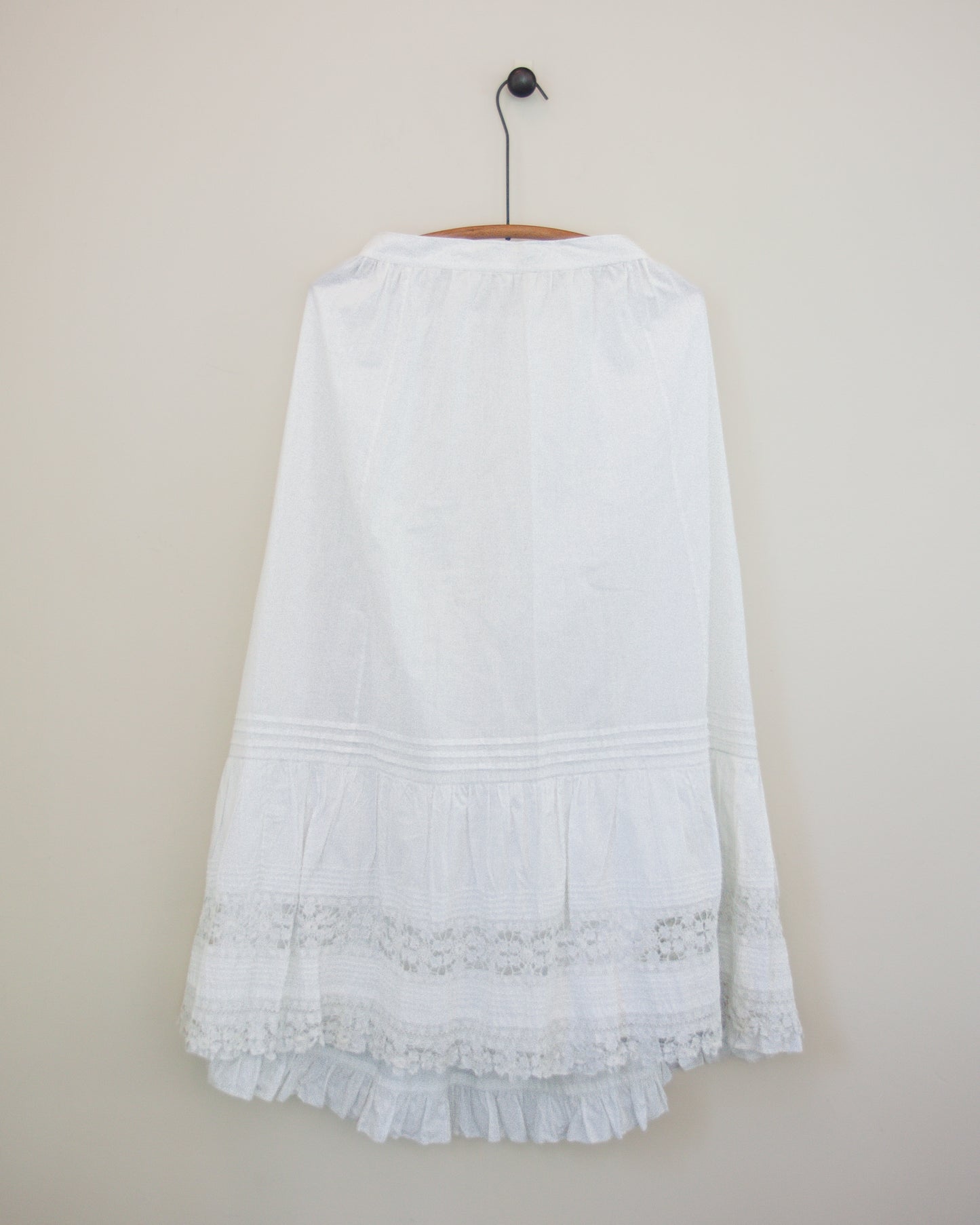 A white skirt with a tiered ruffle hem and lace trim, hanging on a wooden hanger against a plain beige wall.