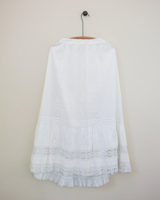 A white skirt with a tiered ruffle hem and lace trim, hanging on a wooden hanger against a plain beige wall.