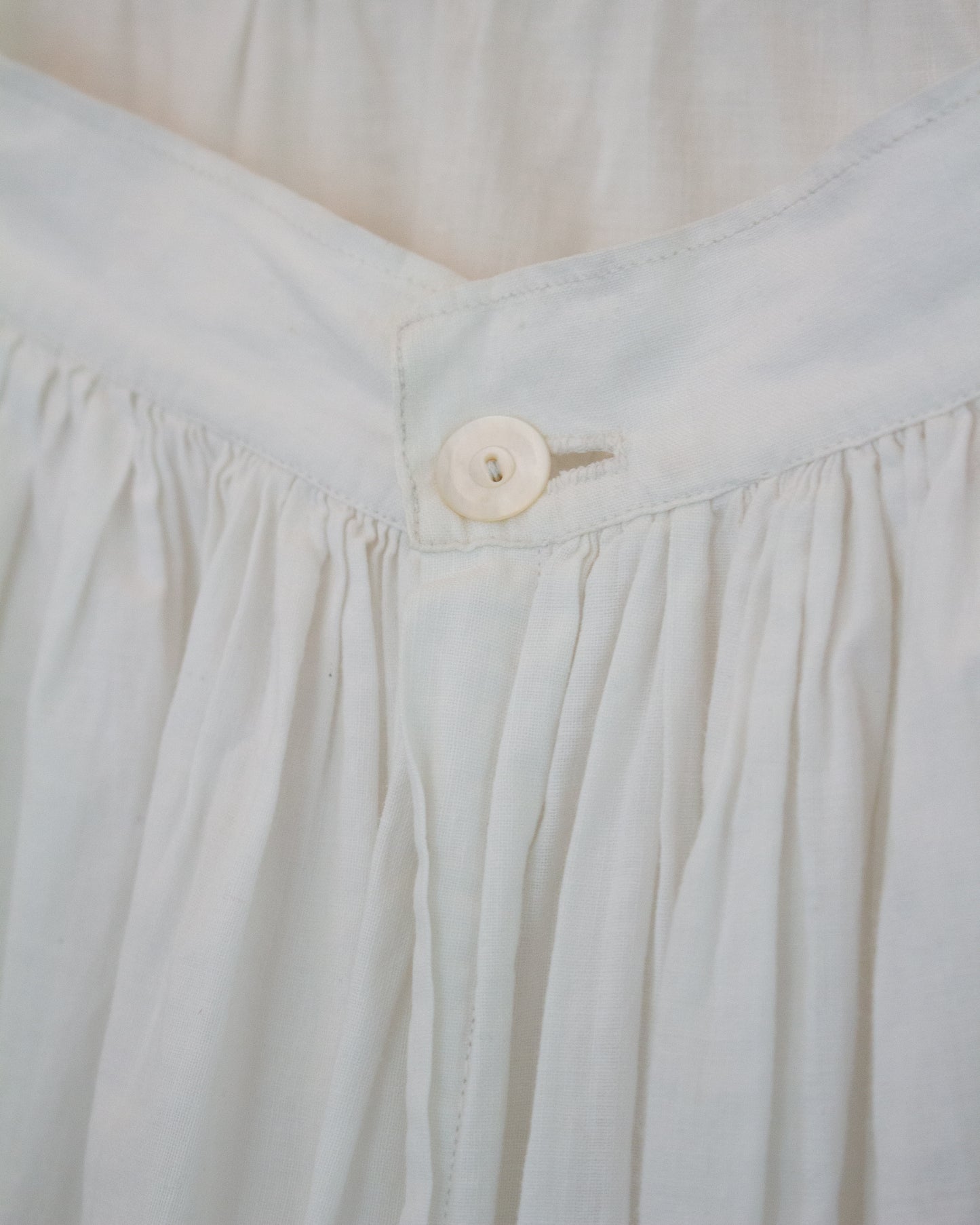 Close-up detail of a white pleated fabric garment, showing gathered seams and a single round button at the waistband.