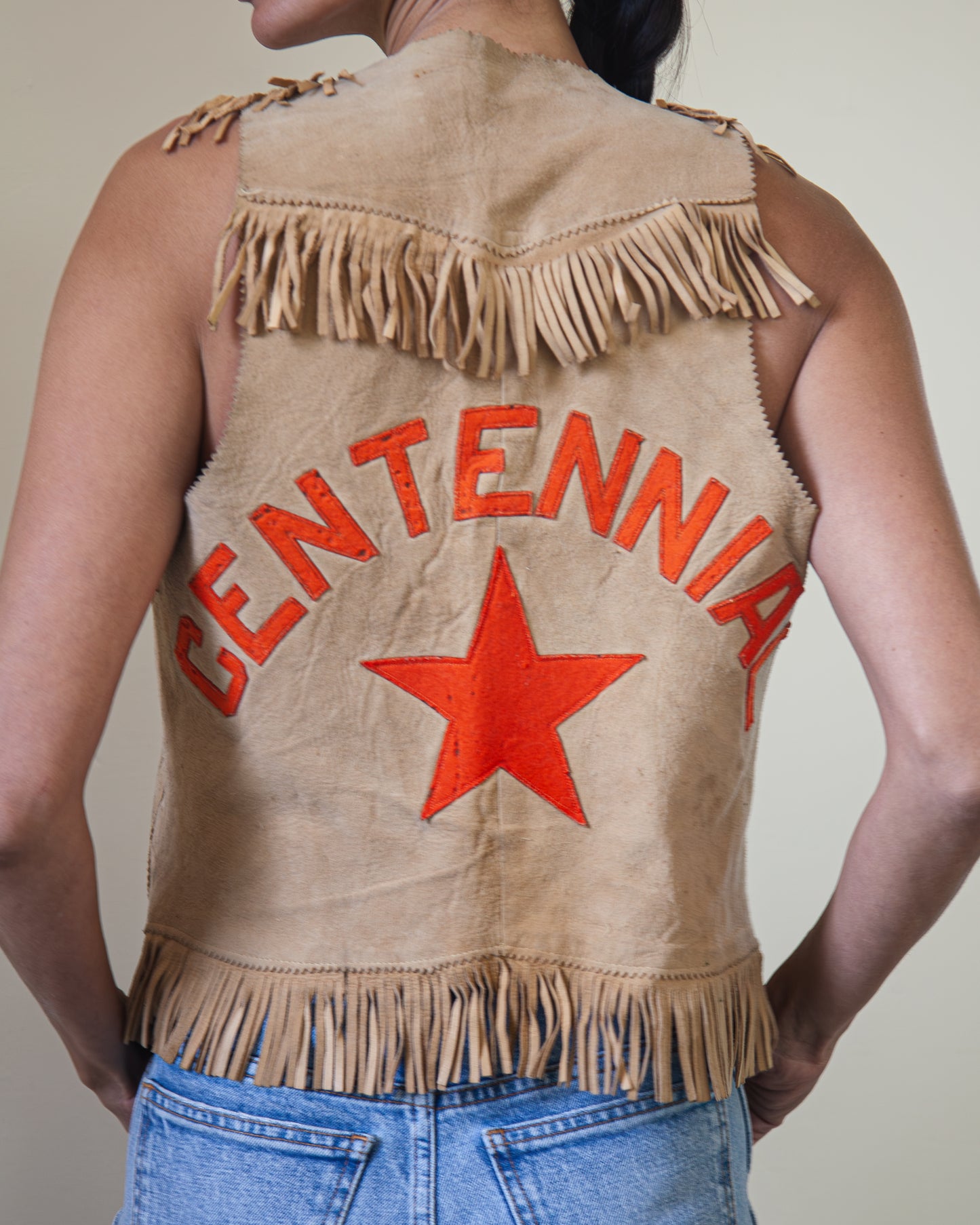 Back view of woman wearing suede leather vest on hanger with orange CENTENNIAL and star graphics on back