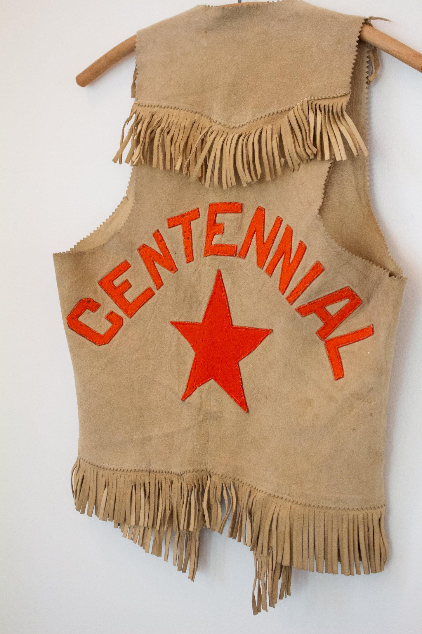 Close up view of back of suede leather vest on hanger with orange CENTENNIAL and star graphics on back