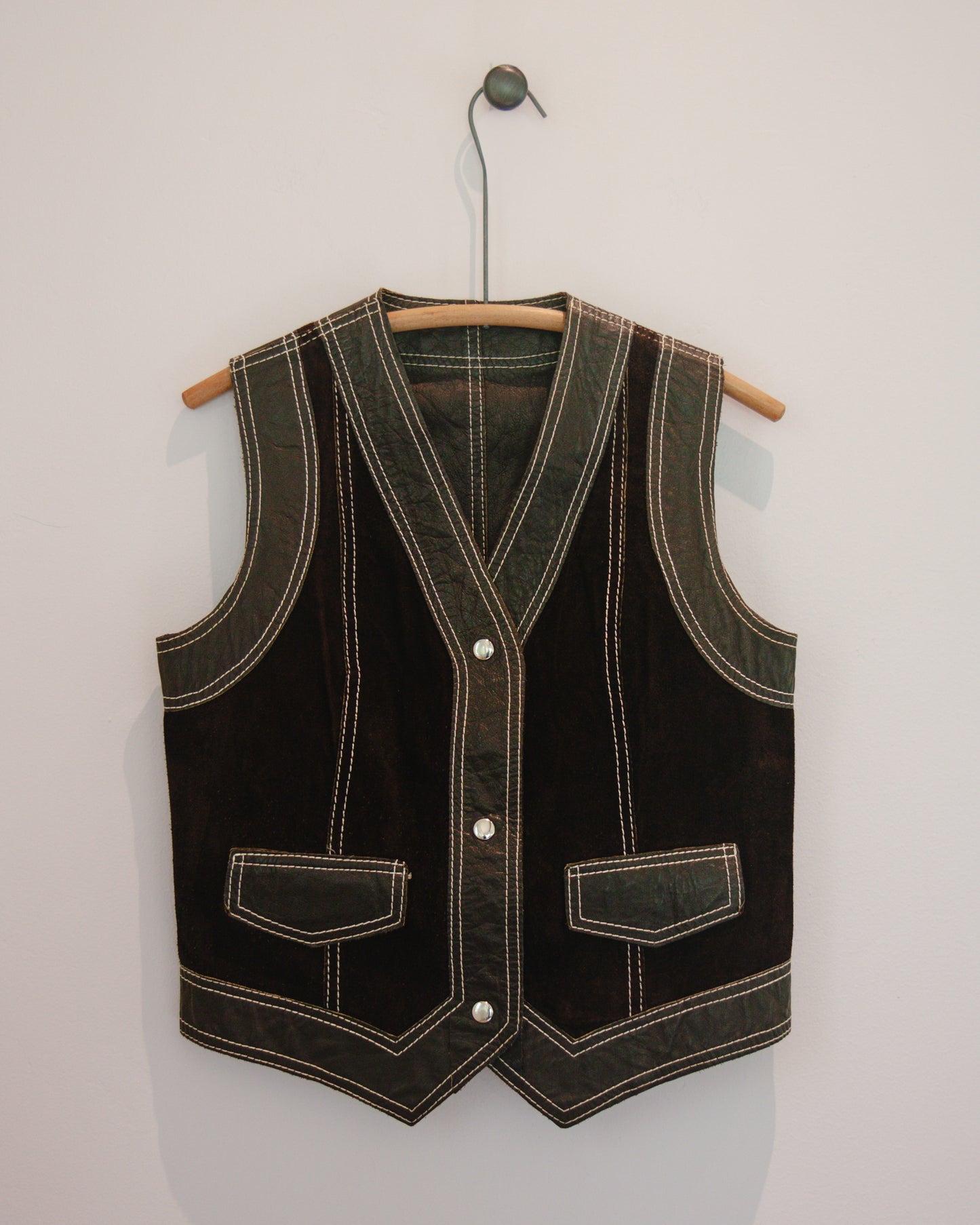Brown leather vest with white stitching on a wooden hanger against a light gray background
