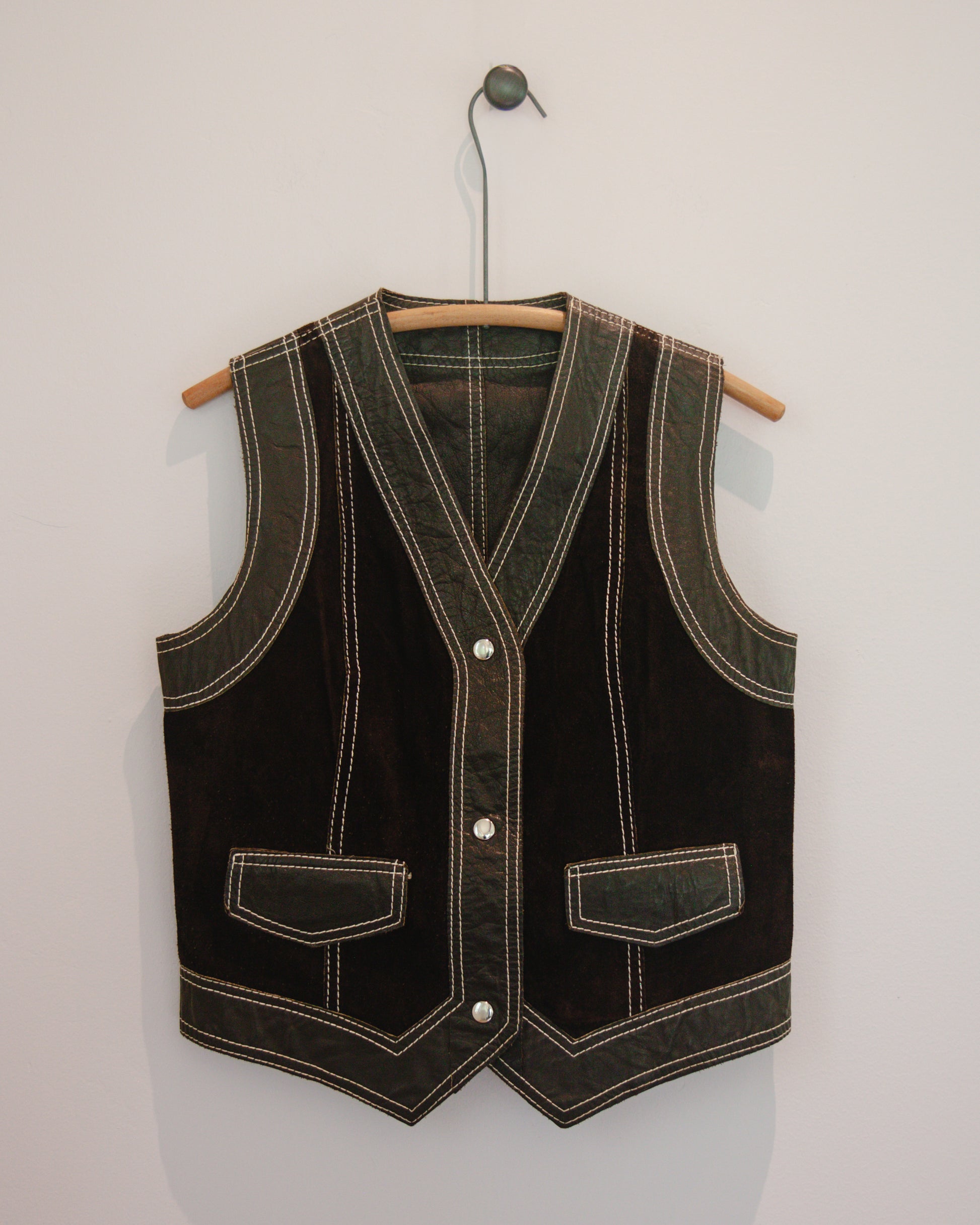 Brown leather vest with white stitching on a wooden hanger against a light gray background