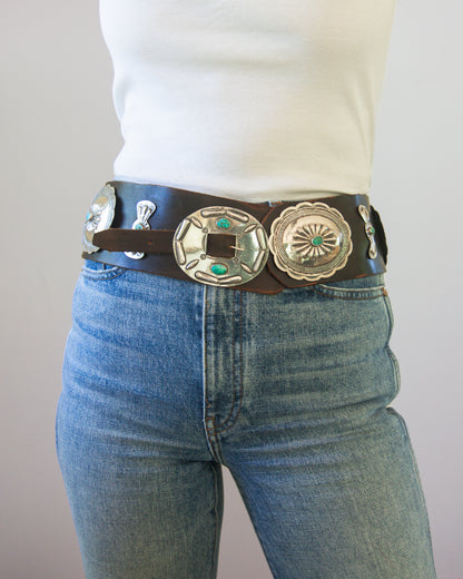 Person wearing blue jeans with a decorative belt featuring large concho-style buckles.