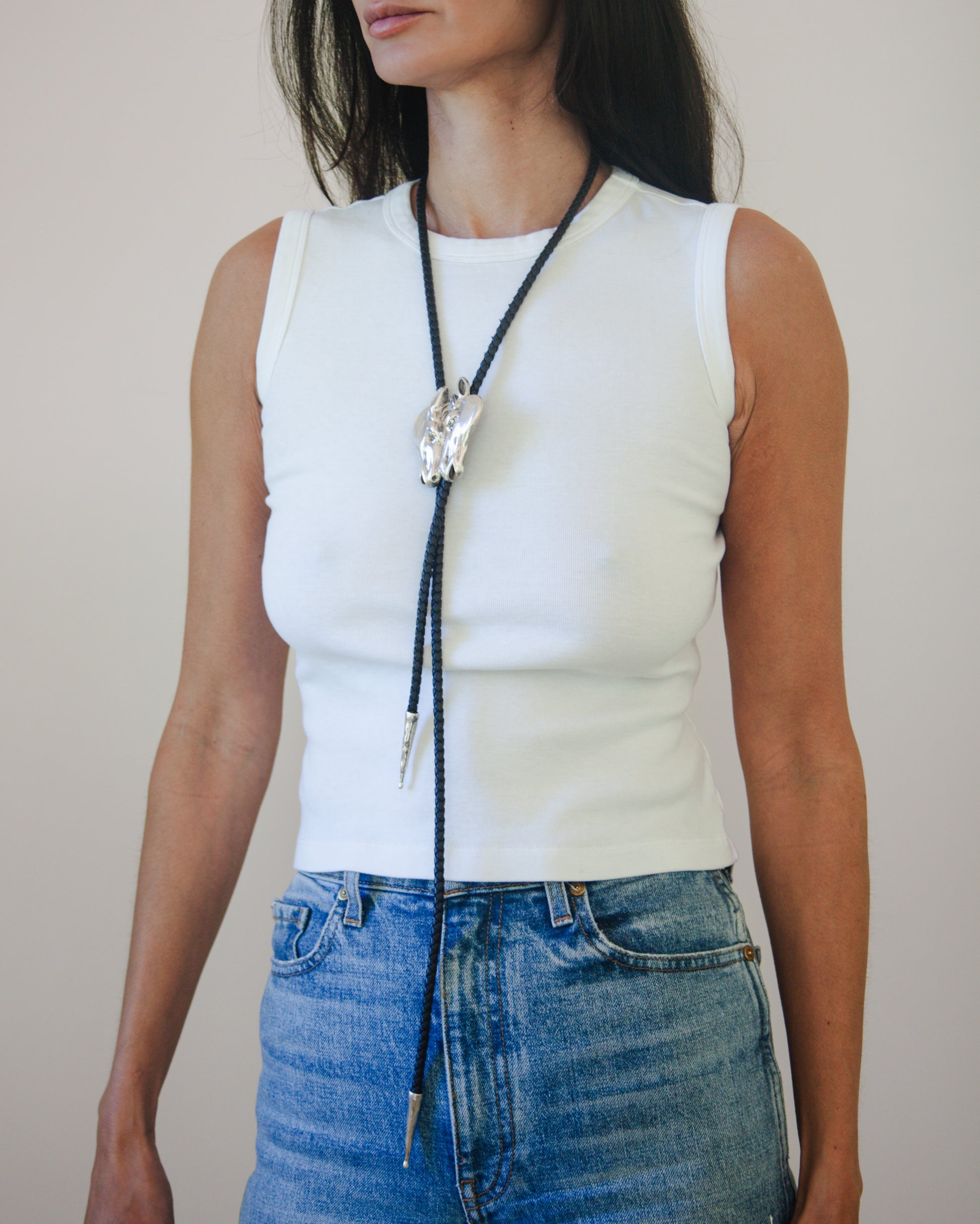 Person wearing a white sleeveless top and blue jeans with a horse head bolo tie