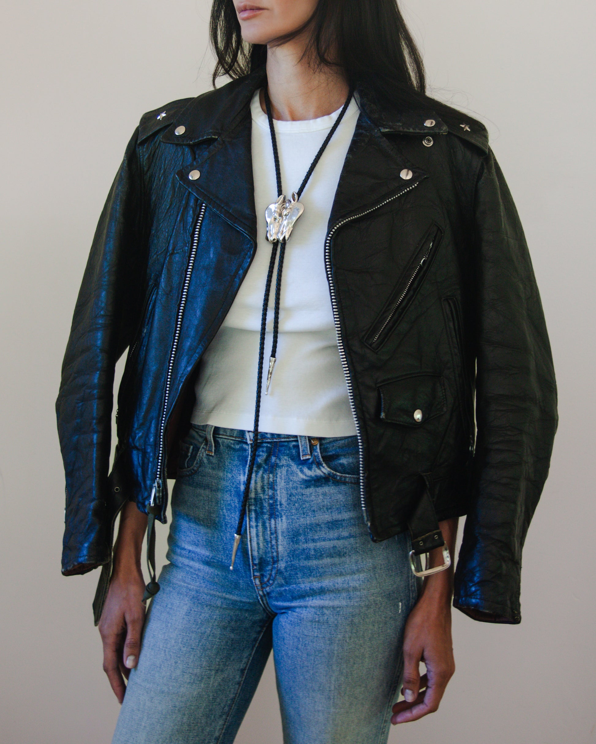 Person wearing a horse head bolo tie with a black leather jacket, white shirt, and blue jeans against a neutral background
