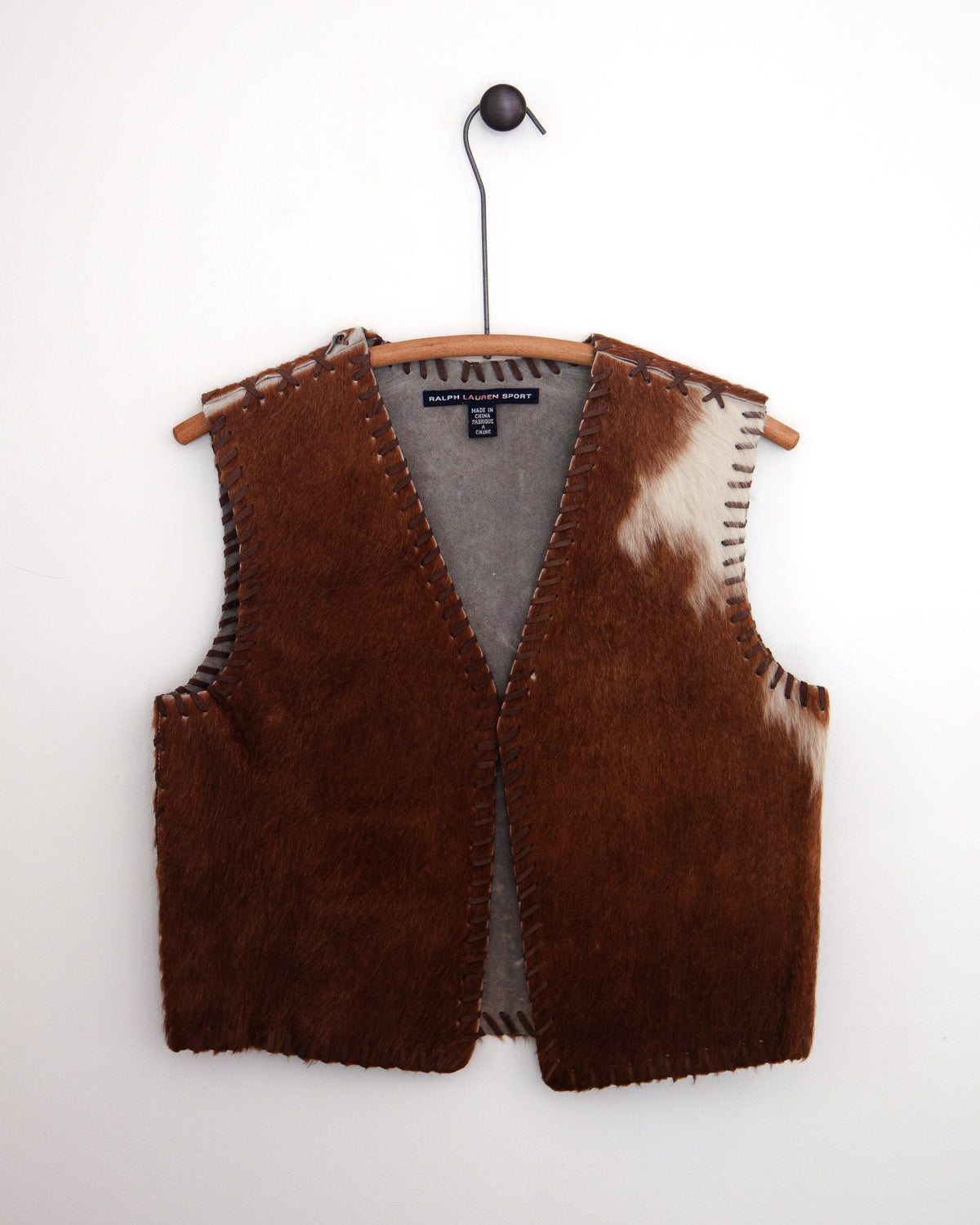 A brown faux fur vest with cowhide accents and whipstitching, hanging on a wooden hanger against a white wall.