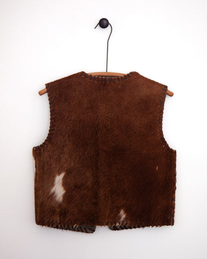 A brown faux fur vest with cowhide accents and whipstitching, hanging on a wooden hanger against a white wall.