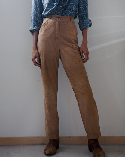 A person wearing high-waisted tan corduroy trousers, a blue patterned button-up shirt, and brown suede shoes, standing against a plain off-white wall.