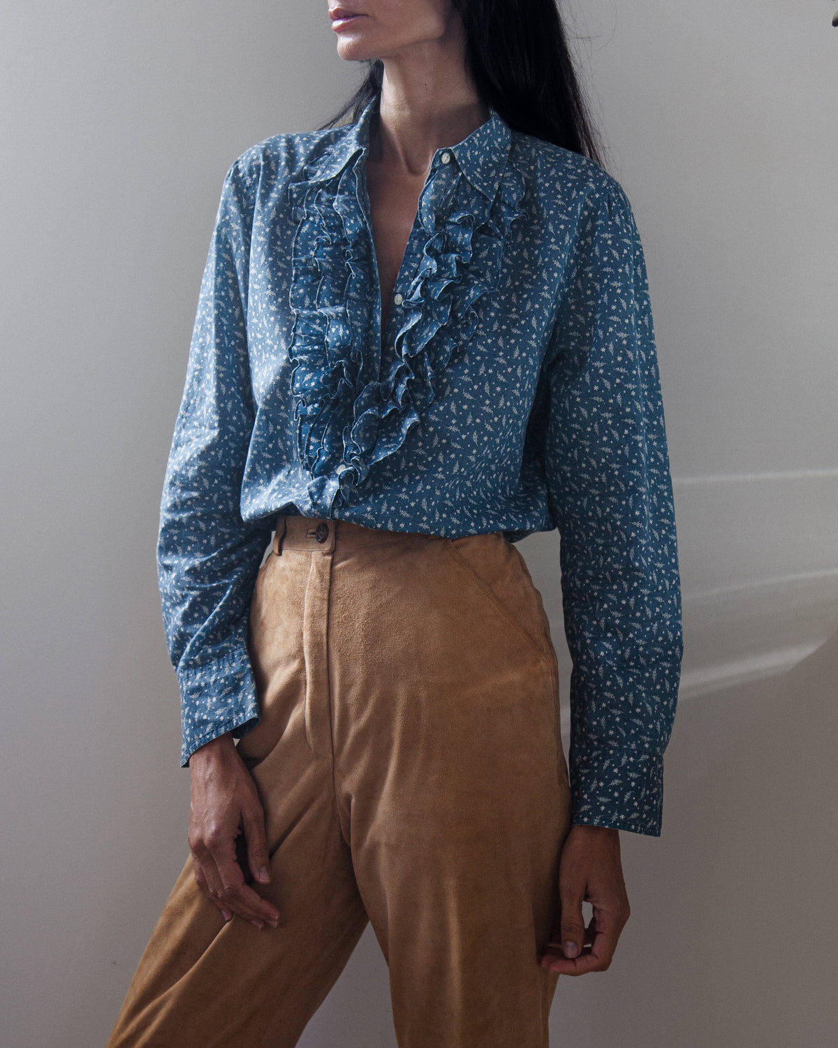 A woman wearing a blue long-sleeve blouse with a white micro-pattern and a prominent ruffle down the front, paired with tan trousers, photographed against a neutral light gray wall.