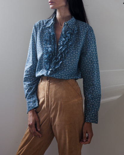 A woman wearing a blue long-sleeve blouse with a white micro-pattern and a prominent ruffle down the front, paired with tan trousers, photographed against a neutral light gray wall.