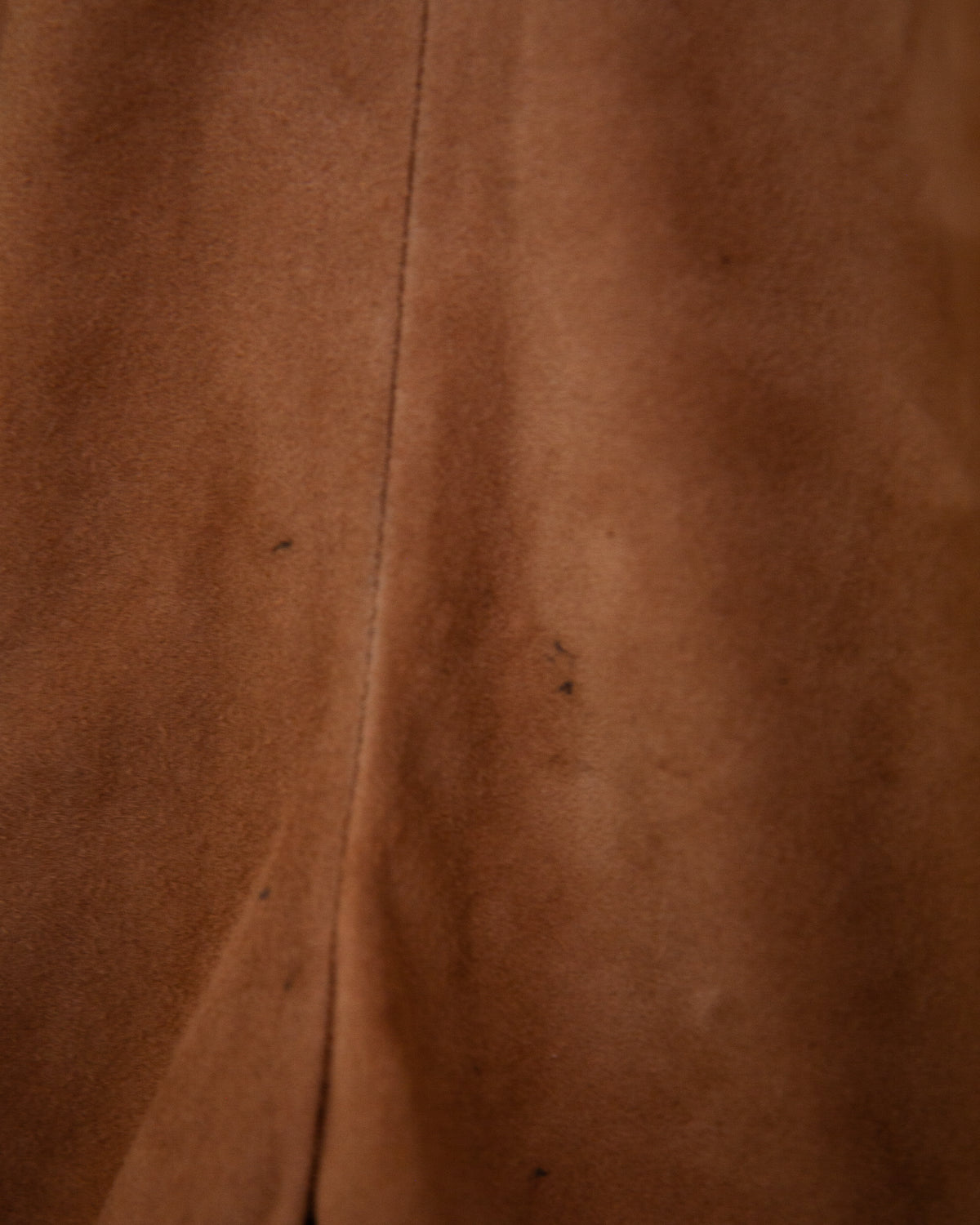 A close-up of a brown suede or suede-like fabric surface with a vertical seam and a few small dark specks.
