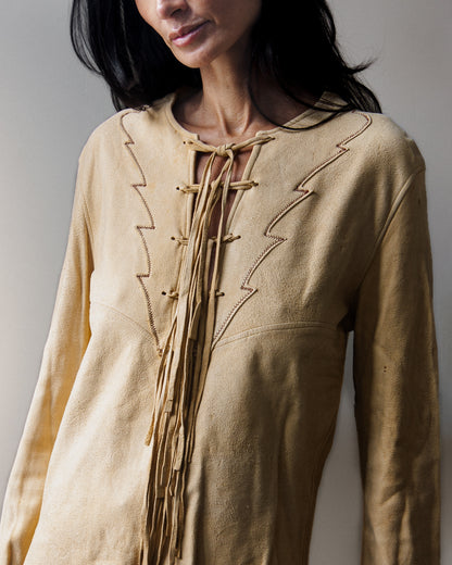 A woman wearing a light beige long-sleeve blouse with decorative tie-front laces and dark blue jeans, standing against a plain light gray wall.