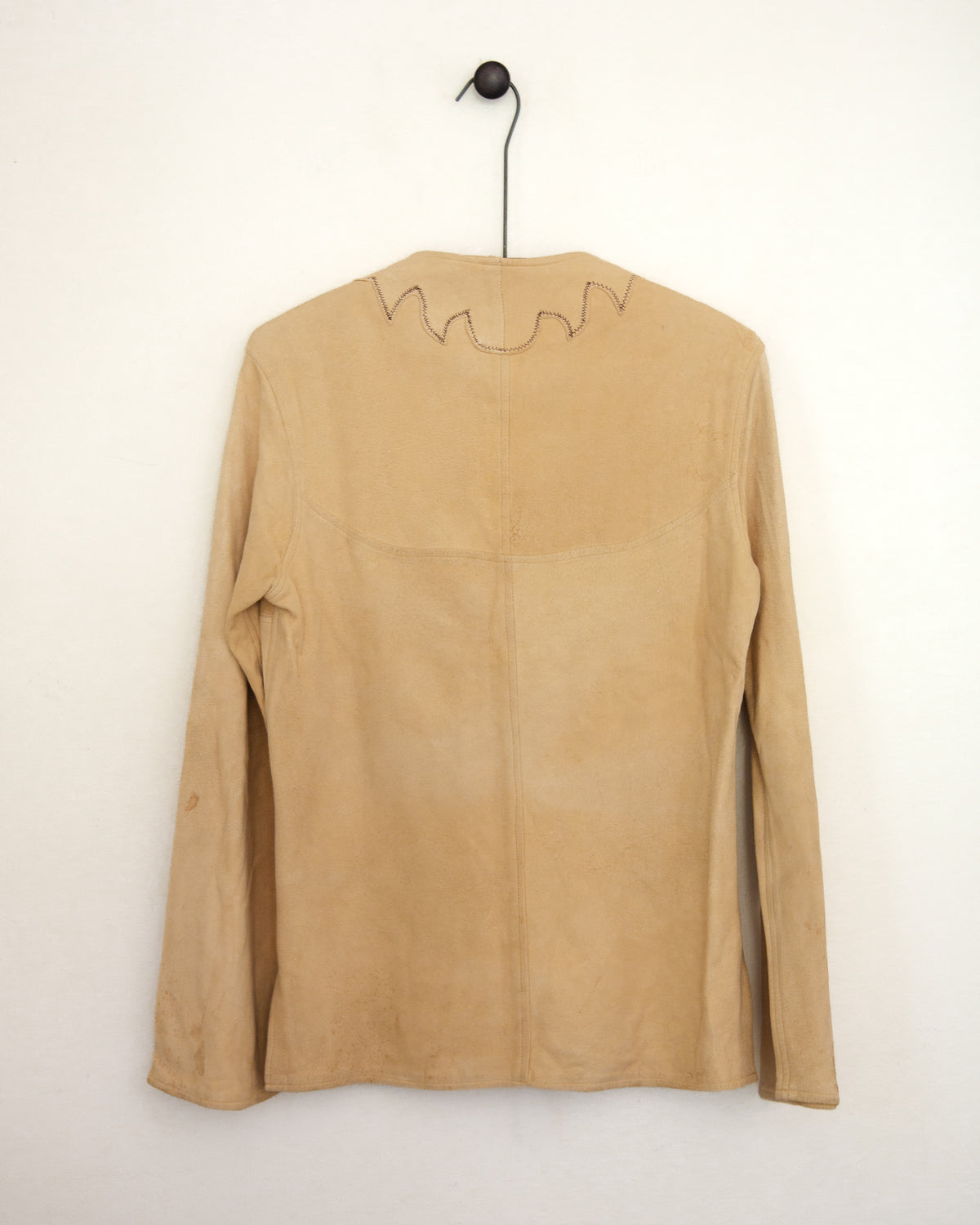 A tan suede long-sleeve top hanging on a black hanger against a plain off-white wall. The back of the garment features a central seam and a jagged, zig-zag stitched detail along the upper neckline.
