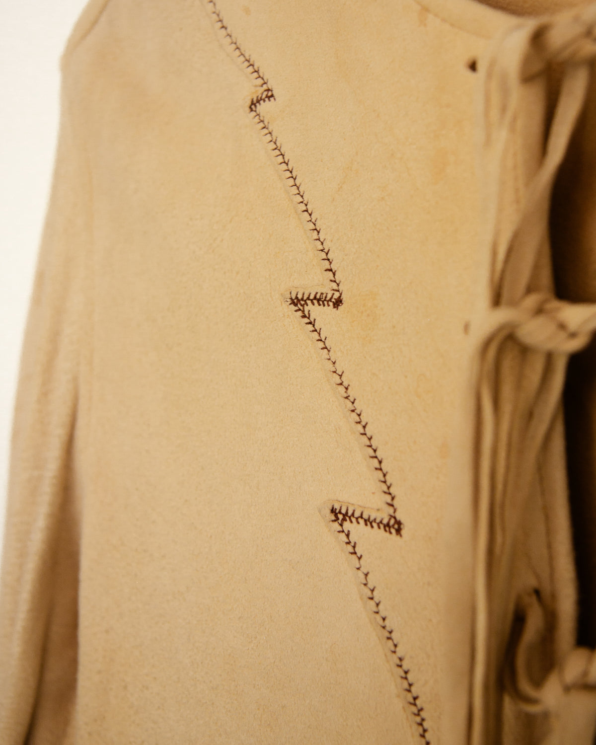 Close-up of a beige suede or leather garment with dark brown zigzag contrast stitching and a matching drawstring detail on the right side.