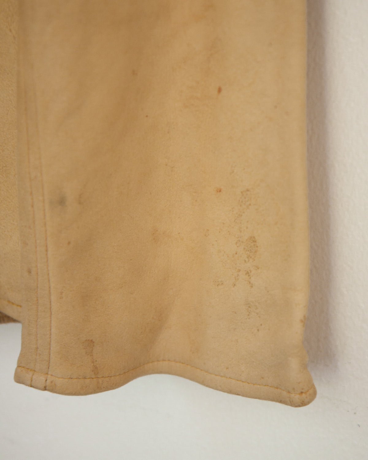 A close-up of a tan suede or leather fabric panel with visible stains and a stitched hem, shown against a plain off-white background.