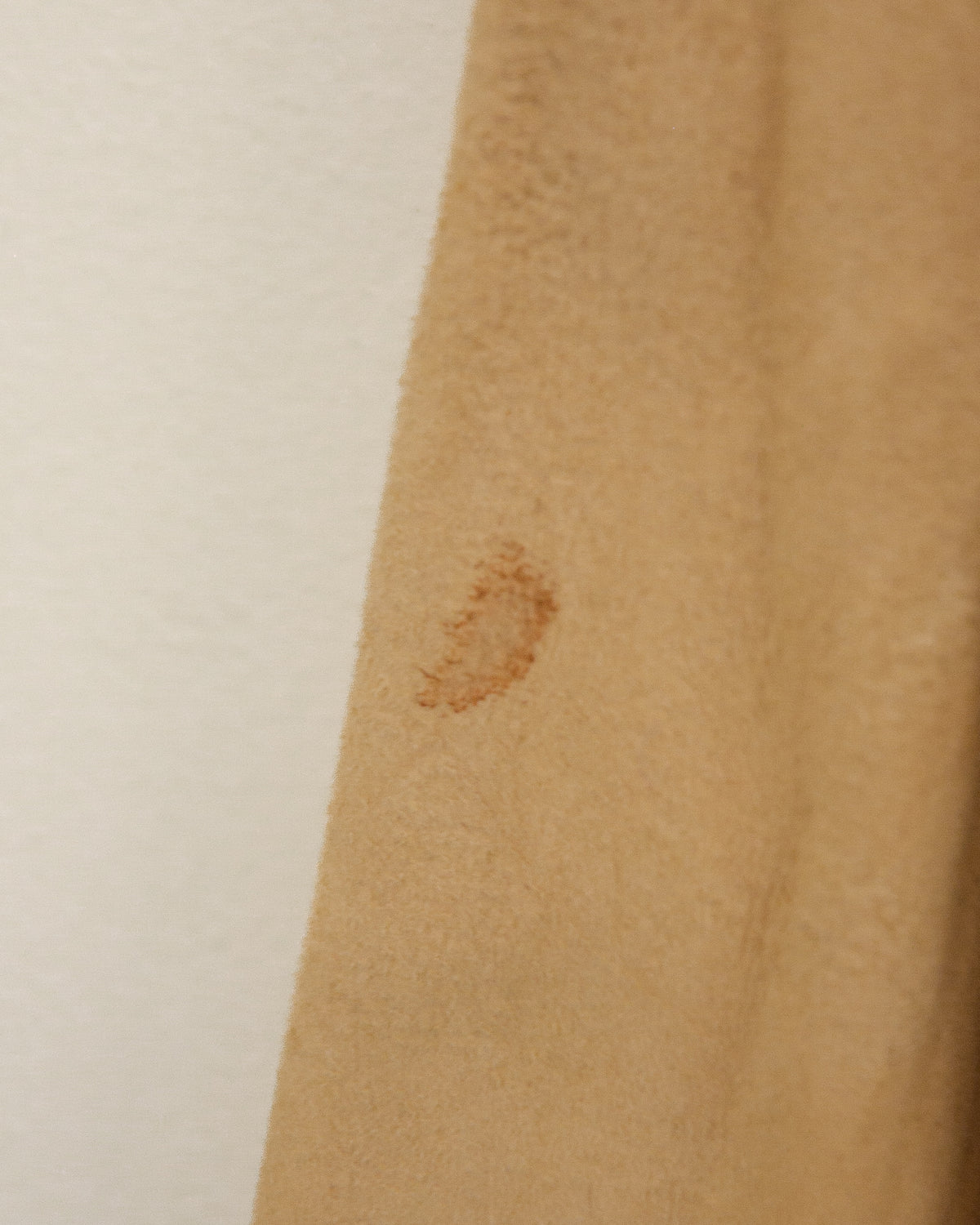 A close-up of a tan suede or leather fabric panel with visible stains and a stitched hem, shown against a plain off-white background.