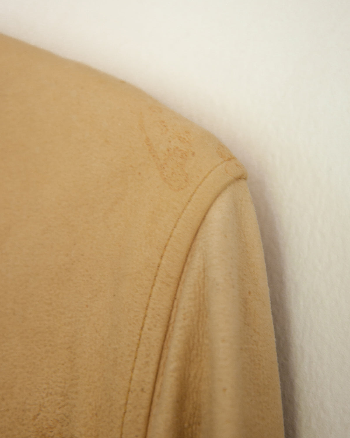 A close-up of a tan suede or leather fabric panel with visible stains and a stitched hem, shown against a plain off-white background.